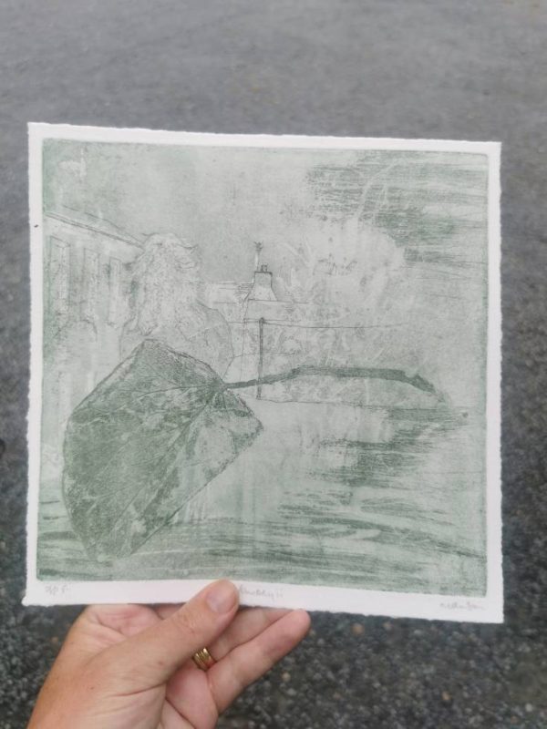 A hand holds a soft green-toned copper plate etching on deckled-edge paper. The print features a large, detailed leaf in the foreground, with the faint outline of a figure and rural rooftops in the background. The texture of the etching and the subtle tonal variations give the piece a dreamlike, atmospheric quality.